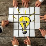 High_angle_view_of_businessmen_hands_touching_white_papers_arranged_on_a_rustic_wooden_table_forming_a_yellow_light_bulb._Conceptual_for_bright_business_ideas_and_innovations._